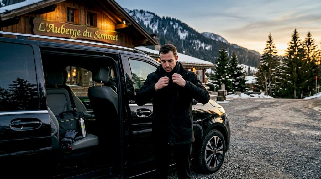 Un chauffeur privé referme les portes d’un minivan dernier cri devant un hôtel de montagne recouvert de neige dans les Alpes.